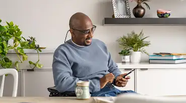 Un hombre en una silla de ruedas con lentes usando un dispositivo Android. De fondo una pared blanca con repisas, libros y plantas. Al frente del hombre una mesa con una taza de café