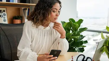 Mujer en una oficina de trabajo usando un dispositivo Android, mirando una laptop y de fondo plantas, ventanas y repisas