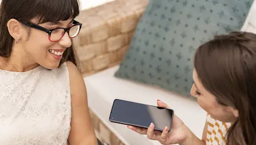 Dos mujeres, una entrevistando a la otra, usando un dispositivo Android para transcribir la entrevista. De fondo un sillón de madera con cojines